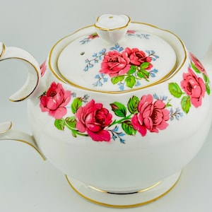 Może przedstawiać: Biały porcelanowy imbryk z pokrywką, uchwytem i dzióbkiem. Imbryk jest ozdobiony różowymi różami, zielonymi liśćmi i niebieskimi akcentami. Złote obramowania zdobią krawędzie imbryka i pokrywki.