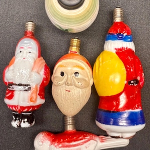 Könnte beinhalten: Eine Sammlung von Vintage-Weihnachtsglühbirnen in verschiedenen Formen. Enthält Weihnachtsmannfiguren in Rot und Weiß, einen Vogel und eine gestreifte Kugel. Die Glühbirnen haben Metallsockel und sind in leuchtenden Farben bemalt.
