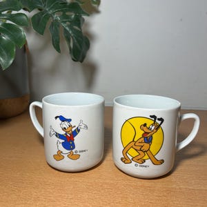 Peut inclure: Deux tasses en céramique blanche avec des personnages Disney. L'une représente Donald Duck en costume de marin bleu, l'autre Pluto dans un cercle jaune. Les deux tasses ont une anse et le logo Disney.