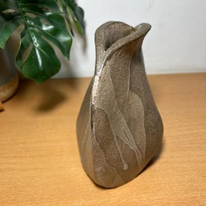 May include: A handcrafted, textured ceramic vase in shades of brown and tan. The vase has a unique, organic shape with a flared opening. The surface features subtle, flowing patterns, adding visual interest to this decorative piece. A stylish addition to any home decor.