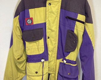 Puede incluir: Una chaqueta de esquí vintage multicolor con un esquema de color amarillo, morado y gris oscuro. La chaqueta tiene cierre de cremallera, múltiples bolsillos y cintura con cordón.