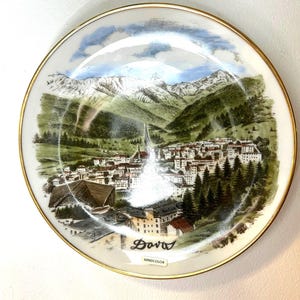 Pode incluir: Um prato de cerâmica branco com uma borda dourada apresenta uma imagem pintada à mão de uma cidade de montanha com picos nevados, árvores verdes e um céu azul. O texto "Davos" está escrito em itálico preto no prato, juntamente com a palavra "Handcolor".
