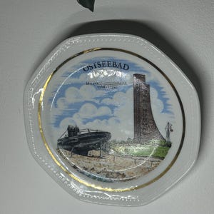 Pode incluir: Um prato de cerâmica branco com uma borda dourada e uma imagem impressa de um submarino e um prédio alto. O prato tem o texto "Ostseebad" impresso nele.