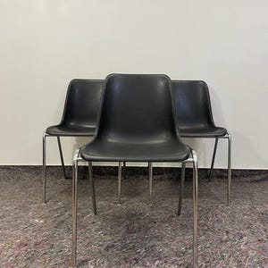 Può includere: Tre sedie nere con gambe cromate. Le sedie hanno un design semplice e moderno con seduta e schienale curvi. Le gambe sono sottili e dritte. Sono davanti a un muro bianco.