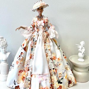 May include: A doll wearing a floral print dress with a matching hat. The dress features a white base with a colorful floral pattern in shades of orange, pink, and brown. The dress has a long skirt and a jacket with lace trim. The doll is wearing a necklace.