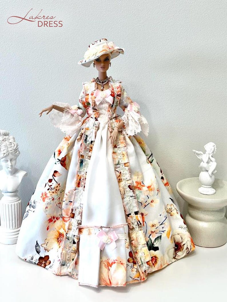 May include: A doll wearing a floral print dress with a matching hat. The dress features a white base with a colorful floral pattern in shades of orange, pink, and brown. The dress has a long skirt and a jacket with lace trim. The doll is wearing a necklace.