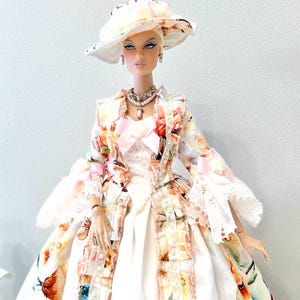 May include: A fashion doll wearing a floral print dress, matching hat, and jewelry. The dress features a white base with colorful floral patterns and lace trim. The hat and jacket have similar floral designs. The doll is accessorized with a pearl necklace and earrings. The text "Lakree Dress" is in the upper left corner.