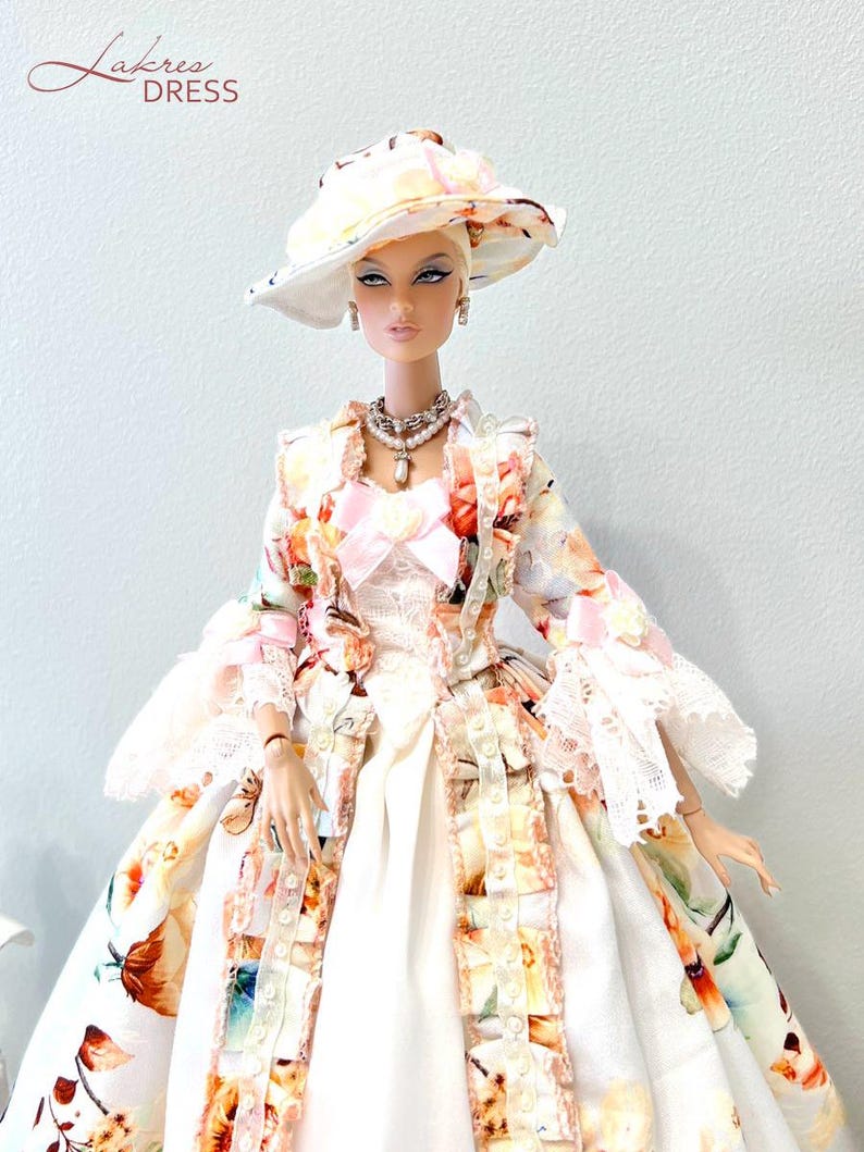 May include: A fashion doll wearing a floral print dress, matching hat, and jewelry. The dress features a white base with colorful floral patterns and lace trim. The hat and jacket have similar floral designs. The doll is accessorized with a pearl necklace and earrings. The text "Lakree Dress" is in the upper left corner.