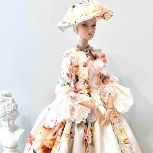 May include: A doll wearing a floral print dress and matching hat. The dress features a white base with orange, pink, and yellow floral patterns. The hat has a wide brim and is adorned with similar floral designs. The doll is accessorized with jewelry.
