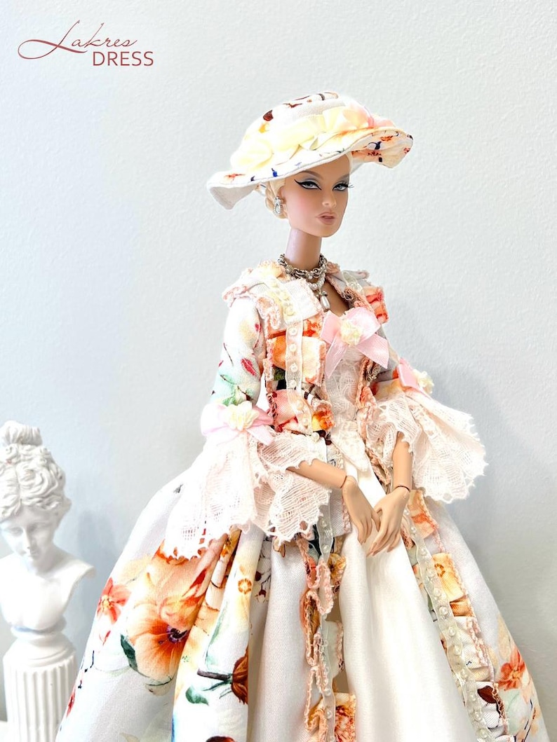May include: A doll wearing a floral print dress and matching hat. The dress features a white base with orange, pink, and yellow floral patterns. The hat has a wide brim and is adorned with similar floral designs. The doll is accessorized with jewelry.