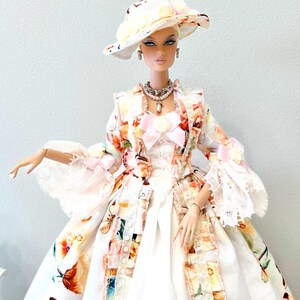 May include: A fashion doll wearing a floral print dress, matching hat, and jewelry. The dress features a white base with colorful floral patterns, lace trim, and a pink bow. The hat has a wide brim with similar floral designs. The doll is posed against a light background.