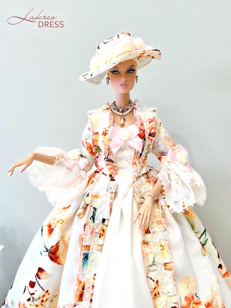 May include: A fashion doll wearing a floral print dress, matching hat, and jewelry. The dress features a white base with colorful floral patterns, lace trim, and a pink bow. The hat has a wide brim with similar floral designs. The doll is posed against a light background.