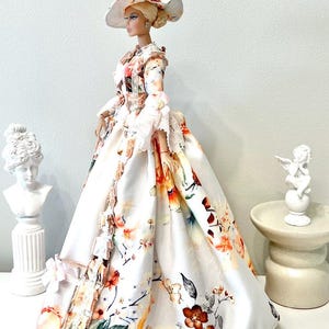 May include: A doll wearing a white floral print dress and matching hat. The dress features a full skirt and long sleeves with lace trim. The hat has a wide brim and is adorned with floral accents. The doll is posed in front of a white background.