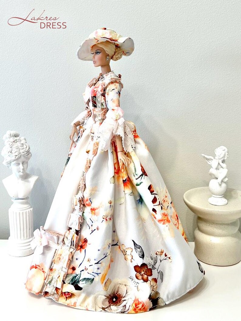 May include: A doll wearing a white floral print dress and matching hat. The dress features a full skirt and long sleeves with lace trim. The hat has a wide brim and is adorned with floral accents. The doll is posed in front of a white background.