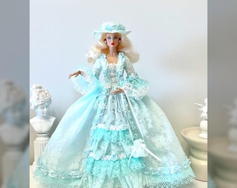 May include: A porcelain doll wearing a light blue and white lace dress with a white hat and pearl earrings. The doll is holding a white lace handbag.