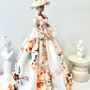 May include: A doll wearing a floral print dress and matching hat. The dress features a white background with orange, pink, and brown floral patterns. The doll is posed with a bust and small statue in the background. The text "Lakree Dress" is in the upper left corner.