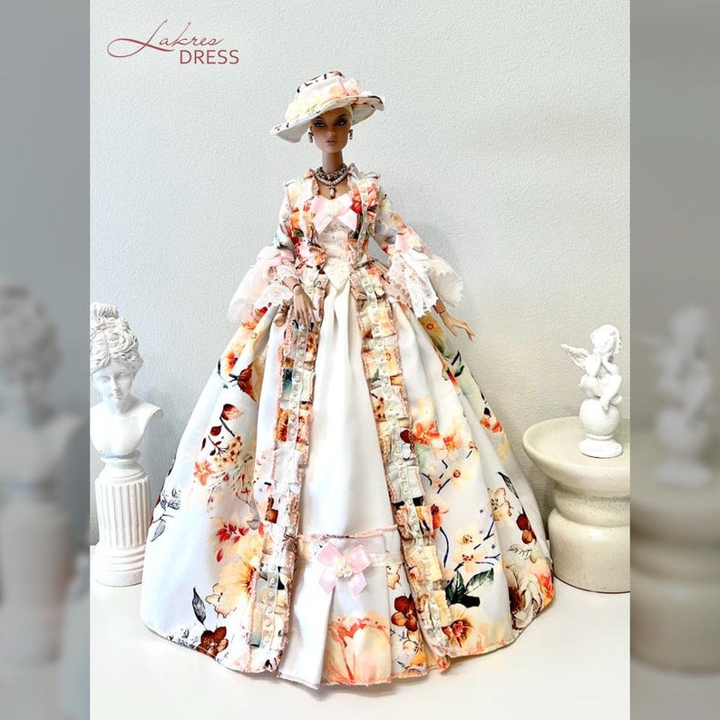 May include: A doll wearing a white and floral patterned dress, hat, and coat. The dress features a full skirt and is adorned with a pink bow and floral accents. The coat has a matching floral pattern and trim. The hat is white with a floral design.