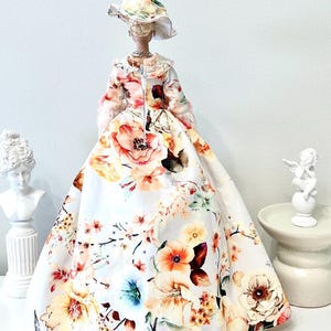 May include: A doll wearing a floral print dress and matching hat. The dress features a full skirt and is adorned with large, colorful flowers in shades of pink, orange, and yellow. The doll is accessorized with a necklace.