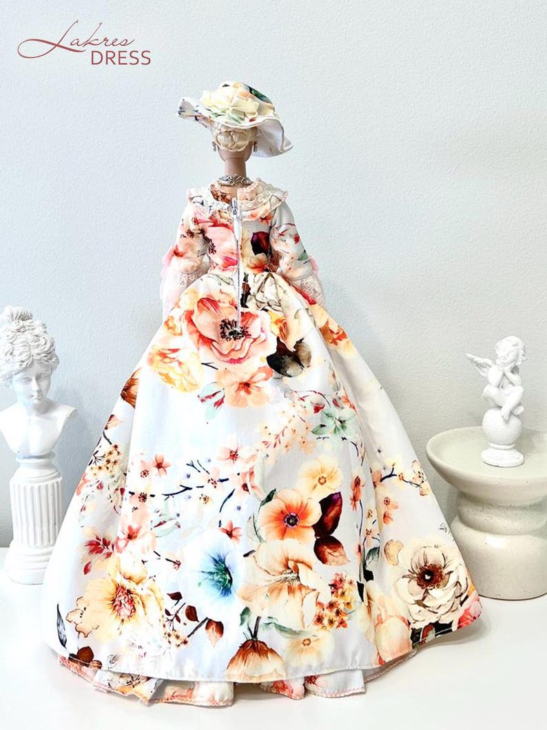 May include: A doll wearing a floral print dress and matching hat. The dress features a full skirt and is adorned with large, colorful flowers in shades of pink, orange, and yellow. The doll is accessorized with a necklace.