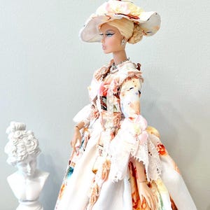 May include: A fashion doll wearing a floral print dress and matching wide-brimmed hat. The dress features a white background with colorful floral patterns and lace trim. The doll has blonde hair and is accessorized with jewelry. The text "Lakree Dress" is in the upper left corner.