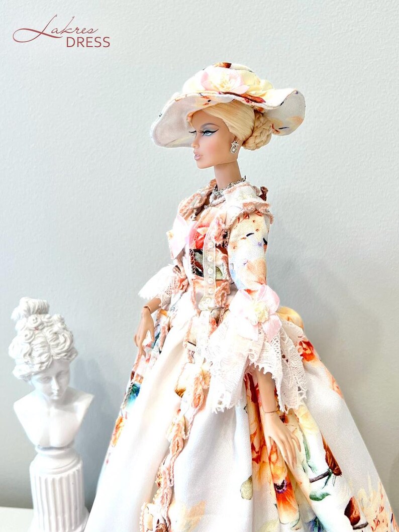 May include: A fashion doll wearing a floral print dress and matching wide-brimmed hat. The dress features a white background with colorful floral patterns and lace trim. The doll has blonde hair and is accessorized with jewelry. The text "Lakree Dress" is in the upper left corner.
