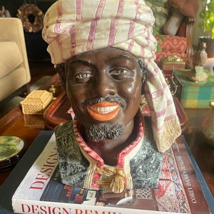May include: A ceramic bust of a person with dark skin, a goatee, and a wide smile, wearing a cream-colored turban with pink stripes. The figure is dressed in a dark green textured garment with a gold-colored decorative clasp at the neckline.