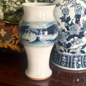 May include: A white ceramic vase with a flared rim and a blue and white painted band around the middle. The vase has a narrow base and a wider, rounded body. The background includes a blue and white patterned vase and greenery.