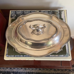 May include: An oval silver-toned serving platter with a matching lid featuring a central handle. The platter has a scalloped, decorative edge and a smooth, reflective metallic finish. The lid is domed with a tiered, stepped design.