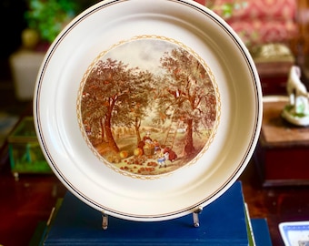 May include: A white ceramic plate with a detailed pastoral scene in the center, featuring trees, figures, and fruit. The plate has a gold trim and a dark brown border. It is displayed on a metal stand, resting on a stack of books.