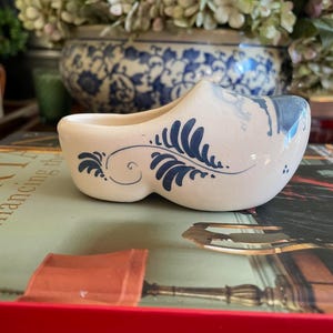 May include: A white ceramic Dutch shoe planter with blue and white Delftware designs. The planter is sitting on a book with the title "Dancing the Plaid" on the cover.
