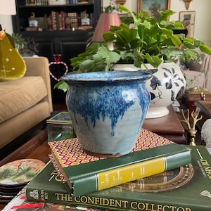 May include: A blue and gray ceramic planter with a textured, dripped glaze. The planter sits atop a stack of books, including one titled "The Confident Collector." The overall theme is home decor and vintage style.