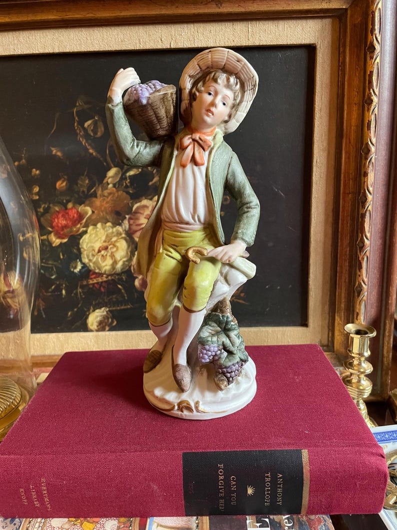 K&ouml;nnte beinhalten: Porzellanfigur eines jungen Mannes mit Hut und gr&uuml;ner Jacke, der einen Korb mit Trauben tr&auml;gt. Die Figur posiert auf einem mit Weinreben bedeckten Sockel, mit roter Fliege und gelber Hose. Die Figur steht auf einem roten Buch.