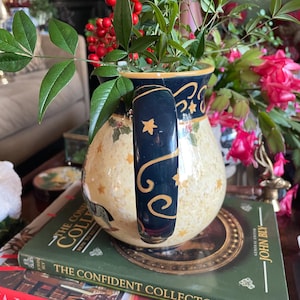 May include: A decorative ceramic pitcher with a navy blue handle and rim, adorned with gold stars and a cream-colored body. The pitcher is filled with greenery and red berries, resting on books.