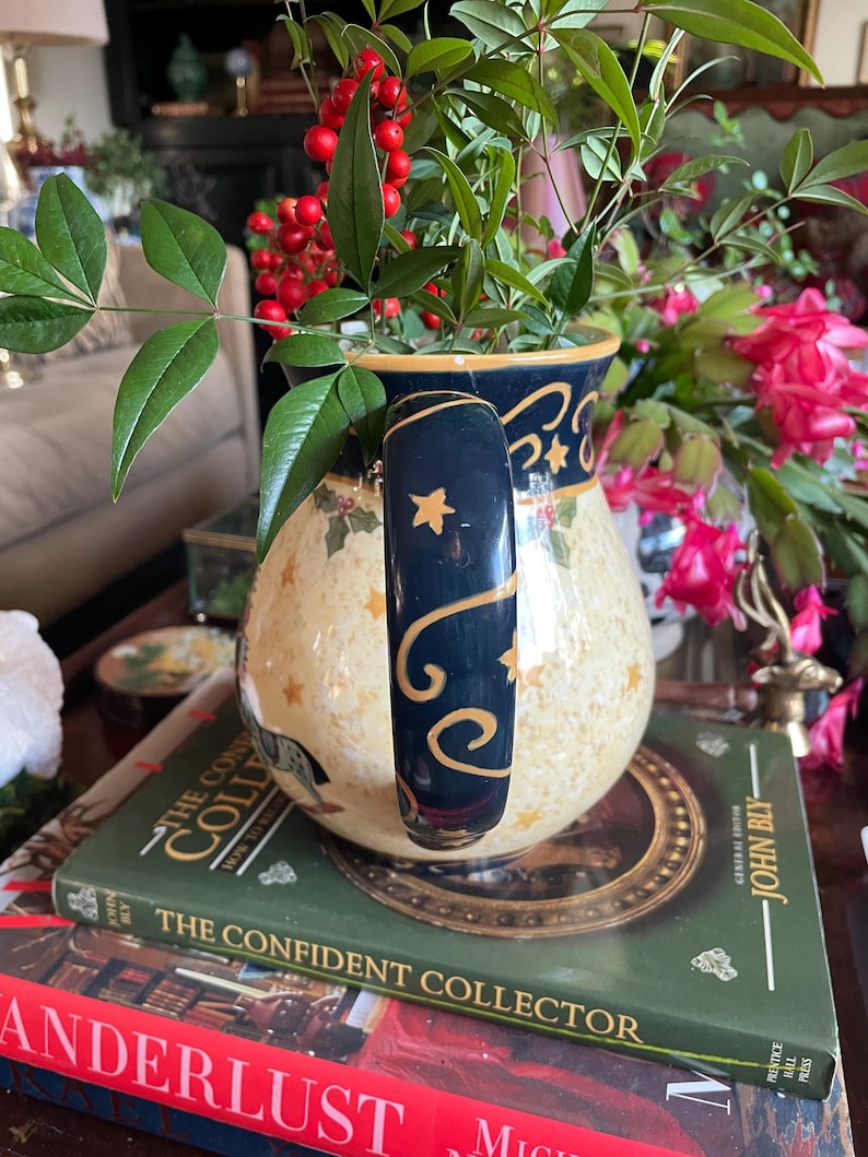 May include: A decorative ceramic pitcher with a navy blue handle and rim, adorned with gold stars and a cream-colored body. The pitcher is filled with greenery and red berries, resting on books.
