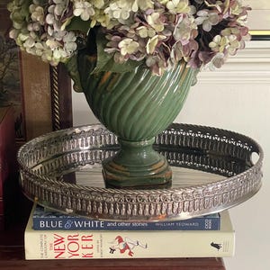Puede incluir: Un jarrón de cerámica verde con un patrón en espiral lleno de flores de hortensia moradas y blancas, sobre una bandeja redonda de metal plateado con un borde ornamentado, colocado sobre dos libros apilados titulados Blue & White y The New Yorker.