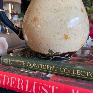 May include: A decorative ceramic pitcher with a cream-colored body, adorned with gold stars and a navy blue handle and rim. The pitcher sits atop a stack of books, including one titled "The Confident Collector."