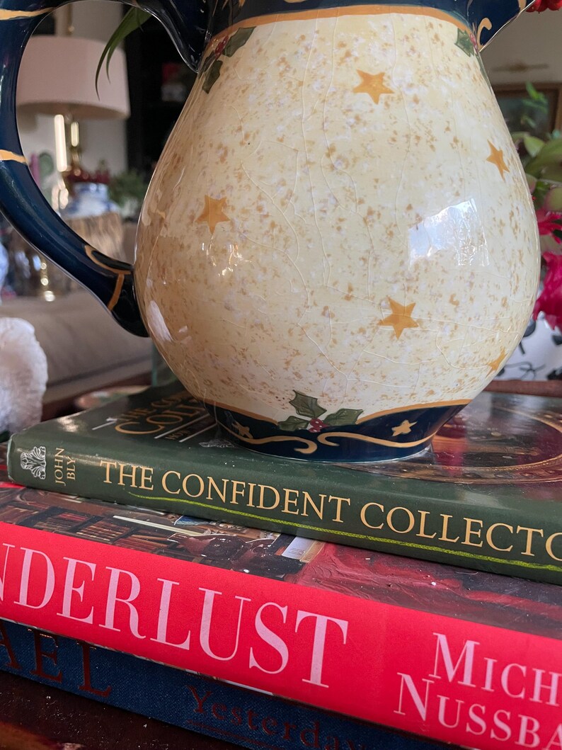 May include: A decorative ceramic pitcher with a cream-colored body, adorned with gold stars and a navy blue handle and rim. The pitcher sits atop a stack of books, including one titled "The Confident Collector."