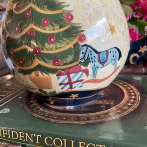 May include: A ceramic Christmas-themed pitcher with a cream-colored background and a detailed illustration of a Christmas tree, ornaments, and a rocking horse. The pitcher has a dark blue handle and base, with gold accents. The design is hand-painted.