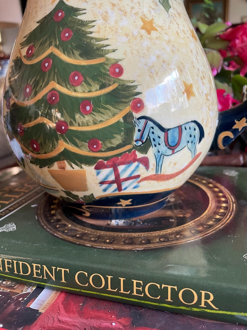 May include: A ceramic Christmas-themed pitcher with a cream-colored background and a detailed illustration of a Christmas tree, ornaments, and a rocking horse. The pitcher has a dark blue handle and base, with gold accents. The design is hand-painted.