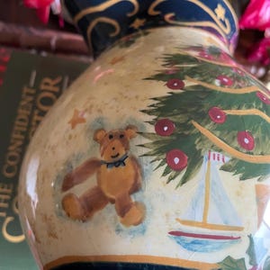 May include: A ceramic pitcher with a Christmas tree and teddy bear design. The pitcher has a cream-colored background with a navy blue rim and base. The Christmas tree is green with red ornaments, and the teddy bear is brown.