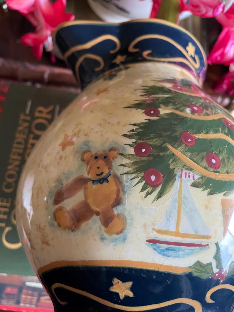 May include: A ceramic pitcher with a Christmas tree and teddy bear design. The pitcher has a cream-colored background with a navy blue rim and base. The Christmas tree is green with red ornaments, and the teddy bear is brown.