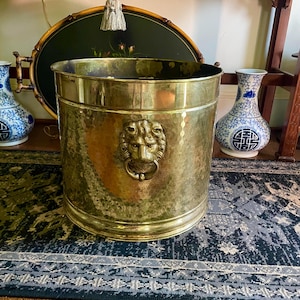 May include: A hammered brass bucket with a lion's head ring pull. The bucket is cylindrical and has a textured surface. The bucket is sitting on a dark blue rug.