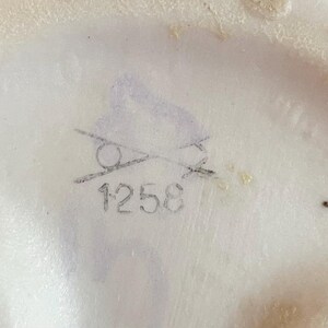 K&ouml;nnte beinhalten: Nahaufnahme einer wei&szlig;en Keramikoberfl&auml;che mit einer verblassten grauen Bleistiftmarkierung. Die Markierung enth&auml;lt einen durchgestrichenen Kreis und die Nummer "1258". Die Oberfl&auml;che hat eine leicht unebene Textur.