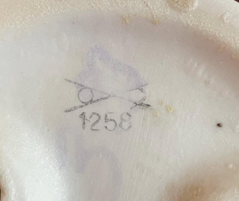 K&ouml;nnte beinhalten: Nahaufnahme einer wei&szlig;en Keramikoberfl&auml;che mit einer verblassten grauen Bleistiftmarkierung. Die Markierung enth&auml;lt einen durchgestrichenen Kreis und die Nummer "1258". Die Oberfl&auml;che hat eine leicht unebene Textur.