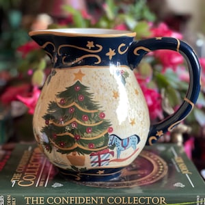 May include: A ceramic pitcher with a Christmas tree design. The pitcher has a cream-colored base with a dark blue rim and handle. The Christmas tree is green with red ornaments, and there are gold stars and a blue rocking horse on the pitcher.