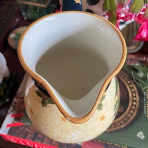 May include: A cream-colored ceramic pitcher with a wide spout and a gold rim. The pitcher features a pattern of gold stars and green holly leaves. The handle is navy blue with a gold accent. The pitcher is sitting on top of a book.