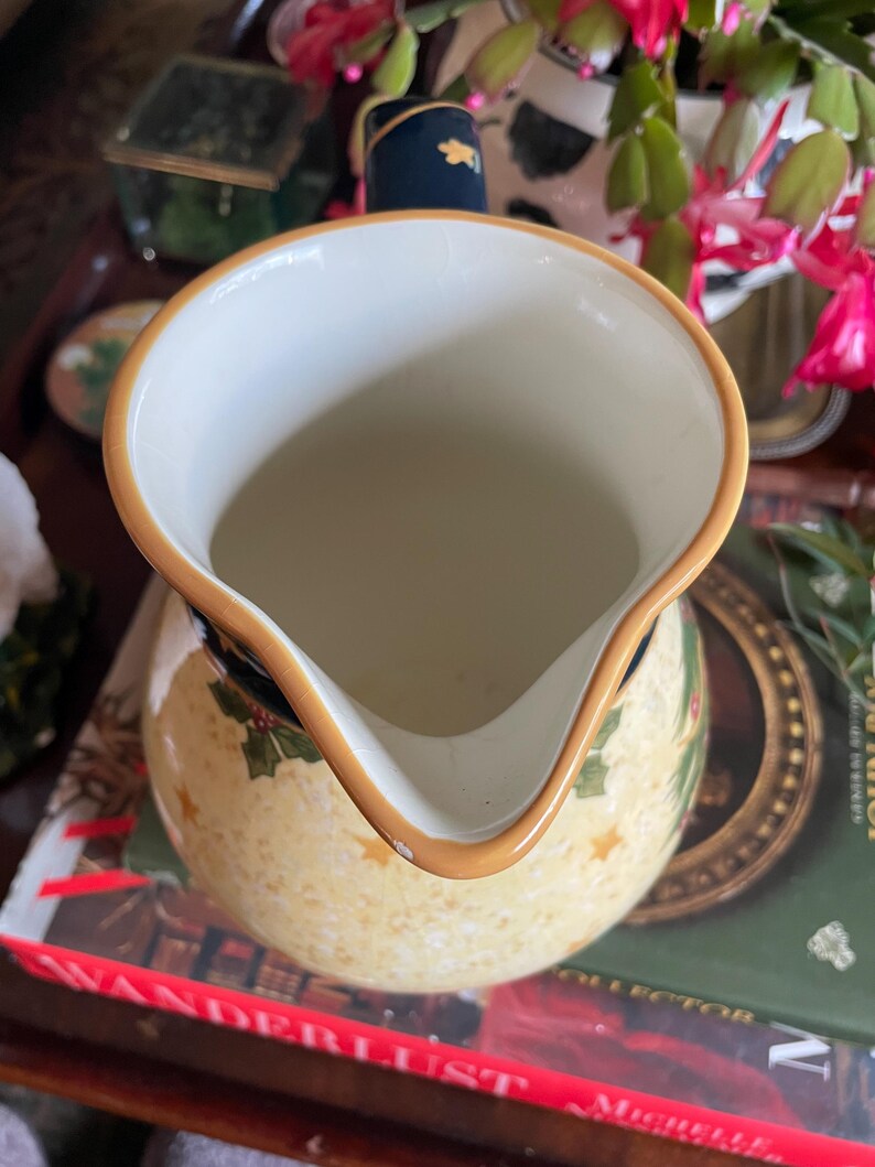 May include: A cream-colored ceramic pitcher with a wide spout and a gold rim. The pitcher features a pattern of gold stars and green holly leaves. The handle is navy blue with a gold accent. The pitcher is sitting on top of a book.