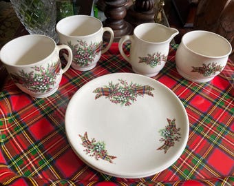 May include: A cream-colored, vintage dish set with a plaid design. The set includes four mugs, a creamer, a sugar bowl, and a plate. Each piece is decorated with a floral and plaid pattern in red, green, and white.
