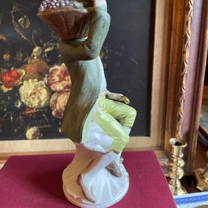 K&ouml;nnte beinhalten: Porzellanfigur eines Mannes, der einen Korb mit Trauben tr&auml;gt. Der Mann tr&auml;gt einen gr&uuml;nen Mantel, gelbe Hosen und braune Schuhe. Die Figur steht auf einem wei&szlig;en Sockel vor einem Gem&auml;lde.