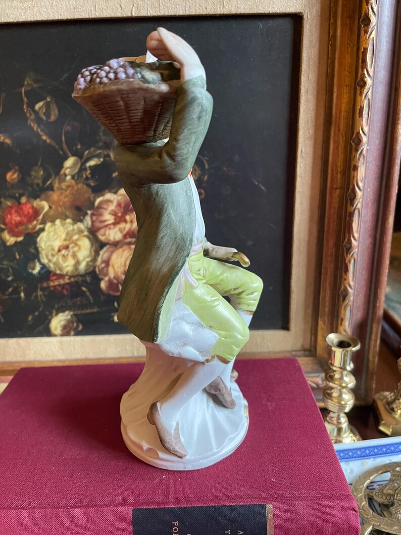 K&ouml;nnte beinhalten: Porzellanfigur eines Mannes, der einen Korb mit Trauben tr&auml;gt. Der Mann tr&auml;gt einen gr&uuml;nen Mantel, gelbe Hosen und braune Schuhe. Die Figur steht auf einem wei&szlig;en Sockel vor einem Gem&auml;lde.